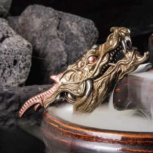 May include: A detailed, golden dragon head incense burner with open jaws and sharp teeth. The dragon's head is positioned above a ceramic bowl, with smoke rising from the bowl. The dragon's head is a detailed sculpture with intricate patterns.