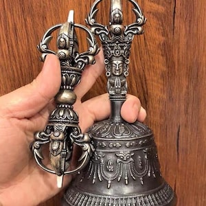 May include: A silver-toned metal vajra and a matching ritual bell. The vajra features intricate symmetrical prongs and a central grip. The bell has a handle topped with a vajra finial, a sculpted face, and a wide base decorated with detailed relief patterns.
