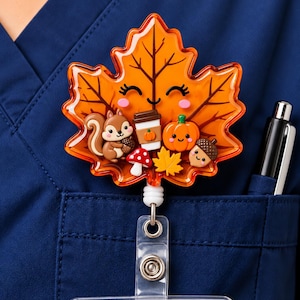 Puede incluir: Un portatarjetas con forma de hoja de arce naranja que presenta una cara sonriente, una ardilla con una bellota, una taza de café, una calabaza, una seta roja y blanca y una pequeña hoja amarilla, sujeto al bolsillo de una casaca azul oscuro.
