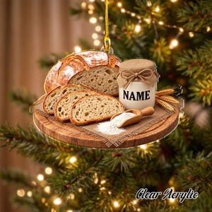 May include: A round hanging ornament featuring a loaf of sourdough bread, three bread slices, a small jar labeled NAME with a burlap lid, a wooden scoop with flour, and wheat stalks on a wooden board, all printed on a clear acrylic surface.
