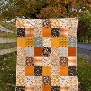 May include: A patchwork quilt featuring a grid of square fabric panels in autumn-inspired colors including orange, brown, cream, and dark gray. The patterns include floral prints, gingham checks, polka dots, and a central square with a squirrel.