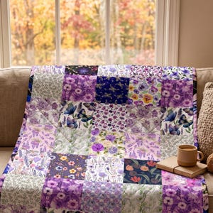 May include: A patchwork quilt featuring a variety of square fabric blocks with purple, lavender, and white floral patterns, butterfly illustrations, and botanical prints. The quilt is draped over a sofa next to a beige ceramic mug and a small book.