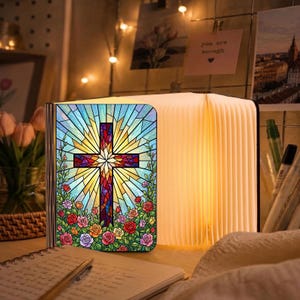 May include: A folding book lamp featuring a stained glass style cover with a central cross design surrounded by colorful roses. The lamp emits a warm, soft glow from its accordion-style pages, creating a decorative light source for a desk.
