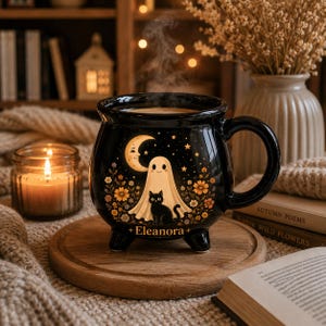 May include: A black ceramic cauldron-shaped mug featuring a graphic of a white ghost, a black cat, a crescent moon, and orange flowers. The name Eleanora is printed in gold text at the bottom. The mug has three small feet and a curved handle.