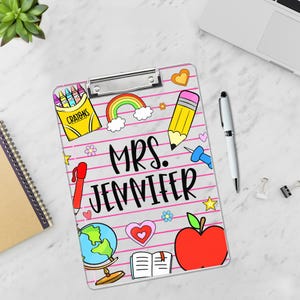 May include: A personalised clipboard featuring the name "MRS. JENNIFER" in black script. The design includes colourful school-themed illustrations such as a rainbow, pencil, apple, globe, and crayons. The background has pink horizontal lines.