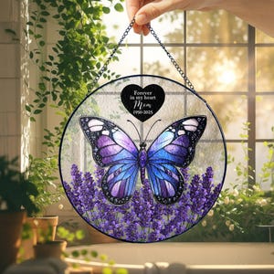 Può includere: Un ornamento rotondo per finestre in vetro colorato con un design a farfalla. La farfalla ha ali blu e viola ed è posta sopra un letto di fiori di lavanda. Il testo "Forever in my heart Mom 1950-2025" è su un cuore nero.