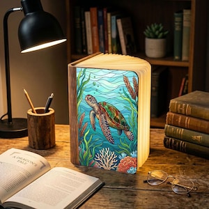 May include: A rectangular book-shaped lamp with a wooden spine and a glowing cover featuring a colorful stained-glass style illustration of a sea turtle swimming among aquatic plants and coral reefs, set on a wooden desk with books and a lamp.