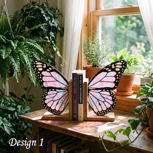 May include: A pair of wooden bookends featuring two butterfly wings with black outlines and iridescent pink panels. The bookends hold two books, The Great Gatsby and To Kill a Mockingbird, upright on a wooden surface. The wings cast light reflections.