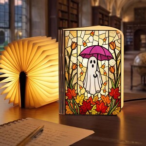 May include: A book-shaped lamp featuring a stained glass design of a white ghost holding a purple umbrella, surrounded by orange tulips, autumn leaves, and candy corn. Beside it, a second book lamp is open, emitting a warm, bright yellow light.