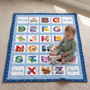 May include: A light blue quilted baby blanket featuring a colourful alphabet grid with animal illustrations for each letter from A to Z. The name Everett is printed in the corners. The border is dark blue with white stars.