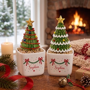 May include: Two square white ceramic planters each containing a decorative Christmas tree and the name Sophia printed in red script. The left tree is green glass with red wire and colorful beads. The right tree is green with white garland.