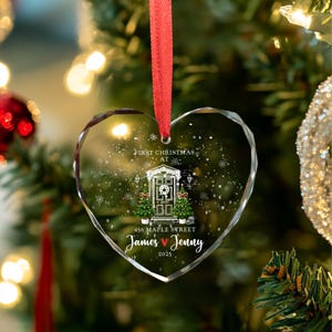 May include: Heart-shaped glass Christmas ornament with a red ribbon. The ornament features a winter scene with a house, Christmas trees, and the text "First Christmas at 456 Maple Street James & Jenny 2025."