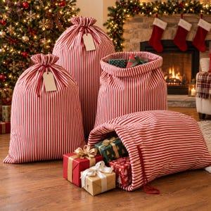 Op de afbeelding: Een collectie grote stoffen cadeautassen met een rood-wit verticaal gestreept patroon. Sommige tassen zijn aan de bovenkant dichtgetrokken met trekkoorden en blanco rechthoekige labels, terwijl andere open zijn en ingepakte cadeaus tonen.
