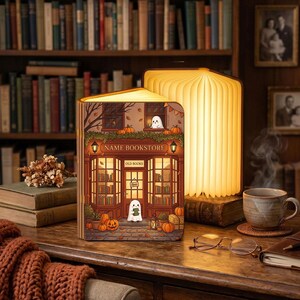 May include: A folding book lamp with a warm glow, featuring a printed illustration of a bookstore storefront titled NAME BOOKSTORE. The scene includes ghosts, pumpkins, and autumn leaves. The lamp is open on a wooden desk next to a mug and glasses.