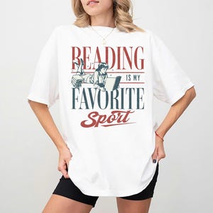 Może przedstawiać: Biała koszulka z napisem "READING IS MY FAVORITE SPORT" w kolorze czerwonym i niebieskim. Grafika przedstawia ilustrację w stylu vintage osoby czytającej. Koszulka ma krótkie rękawy i luźny krój.