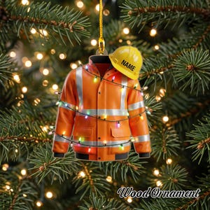 May include: A flat, hanging ornament shaped like an orange high-visibility jacket with silver reflective stripes and a yellow hard hat resting on the shoulder. The hard hat features the text NAME. A string of multicolored Christmas lights wraps around.