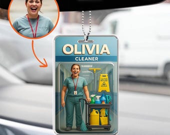 Op de afbeelding: Een auto luchtverfrisser met de naam "OLIVIA CLEANER". Het ontwerp toont een foto van een vrouw in een teal uniform, een schoonmaakwagen en een "Pas op natte vloer" bord. Het hangt aan een zilveren ketting.