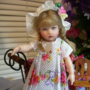 May include: A porcelain doll with blonde hair wearing a white dress with black polka dots and pink flowers. The doll is wearing a white bonnet with a pink flower and a ribbon. The doll is holding a brown wooden handle.