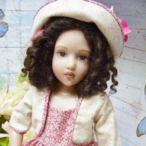 May include: A porcelain doll wearing a pink floral dress with a white cardigan and a white hat with pink trim. The doll has brown curly hair and brown eyes.