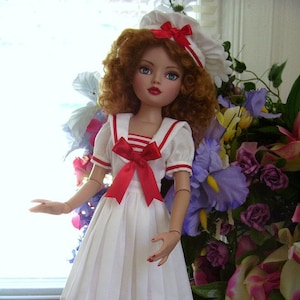 Może przedstawiać: Porcelanowa lalka w białej sukience marynarskiej z czerwonymi lamówkami i czerwoną kokardką. Lalka ma rude włosy, niebieskie oczy i czerwoną szminkę. Lalka stoi przed bukietem kwiatów.