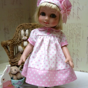 Può includere: Bambola vintage con un vestito a pois rosa e bianco, cappello abbinato e scarpe bianche. La bambola ha i capelli biondi ed è posizionata davanti a una piccola sedia di vimini. Sono visibili anche un coniglietto giocattolo e un orsacchiotto.