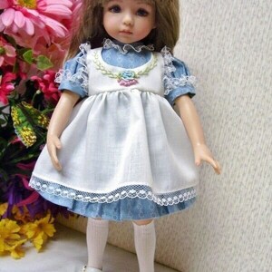 May include: A porcelain doll with brown hair and blue eyes, wearing a blue and white dress with lace trim. The doll is standing, wearing white shoes and knee-high socks. A floral arrangement is in the background.