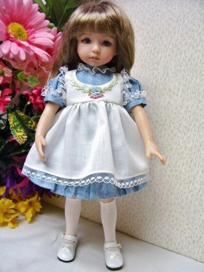 May include: A porcelain doll with brown hair and blue eyes, wearing a blue and white dress with lace trim. The doll is standing, wearing white shoes and knee-high socks. A floral arrangement is in the background.