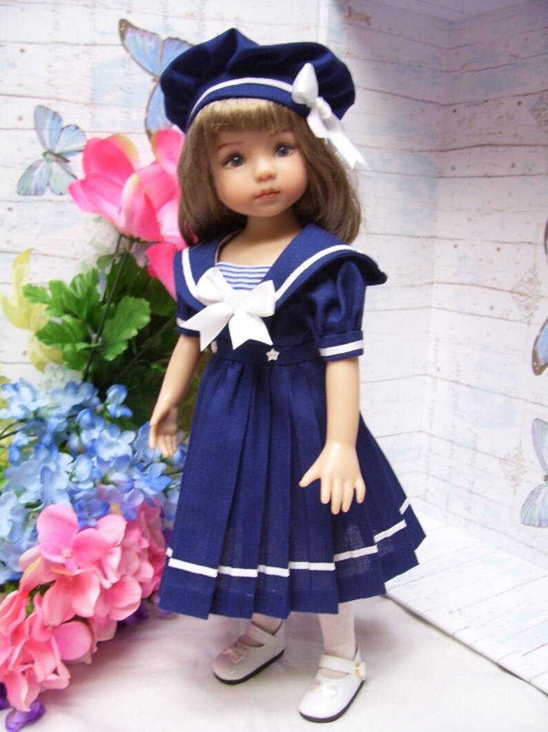 May include: A doll wearing a blue sailor dress with white trim and a white bow. The doll has brown hair and blue eyes and is wearing a blue and white sailor hat.