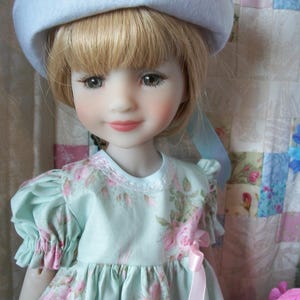 May include: A porcelain doll with blonde hair and blue eyes, wearing a pale blue dress with pink rose patterns and a matching hat. The dress has short, puffed sleeves and a pink ribbon. The doll is posed in front of a floral quilt.