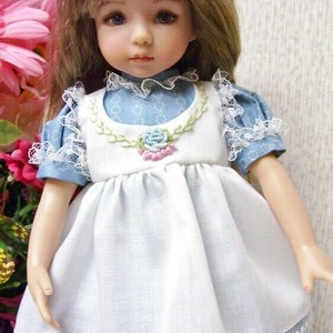 May include: A porcelain doll with blonde hair and blue eyes, wearing a white dress with a blue floral print and lace trim. The dress has a white bib with a floral embroidery and a blue collar. The doll is posed in front of a pink flower arrangement.