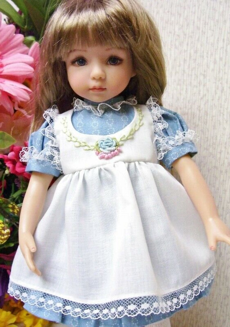 May include: A porcelain doll with blonde hair and blue eyes, wearing a white dress with a blue floral print and lace trim. The dress has a white bib with a floral embroidery and a blue collar. The doll is posed in front of a pink flower arrangement.