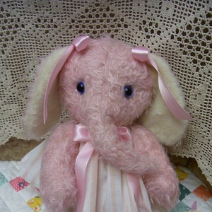 May include: A pink plush elephant toy with white ears and a pink satin ribbon around its neck. The elephant has blue eyes and a long, curly trunk.