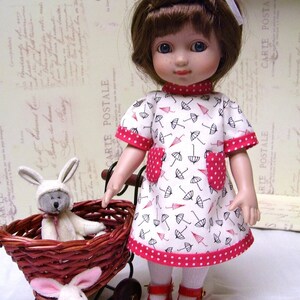 May include: A doll wearing a white dress with a red polka dot trim and black umbrella print. The doll has brown hair and blue eyes. The doll is holding a wicker basket with two stuffed bunny rabbits.