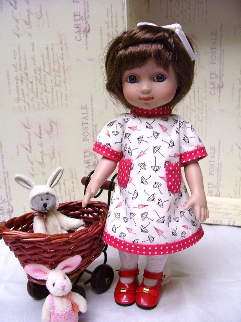 May include: A doll wearing a white dress with a red polka dot trim and black umbrella print. The doll has brown hair and blue eyes. The doll is holding a wicker basket with two stuffed bunny rabbits.