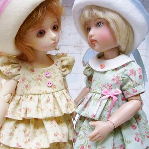 May include: Two dolls wearing floral print dresses and hats. The doll on the left is wearing a white hat with a pink ribbon and a yellow dress with a floral pattern. The doll on the right is wearing a white hat with a blue ribbon and a green dress with a floral pattern.