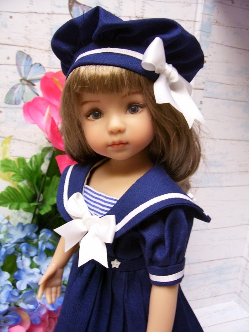 May include: A doll wearing a navy blue sailor dress with white trim and a white bow. The doll has brown hair and blue eyes and is wearing a navy blue beret with a white ribbon.