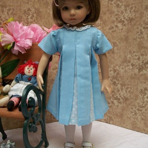 May include: A blue and white polka dot dress with a white lace collar and a pink bow in the hair. The doll is wearing white tights and white shoes.