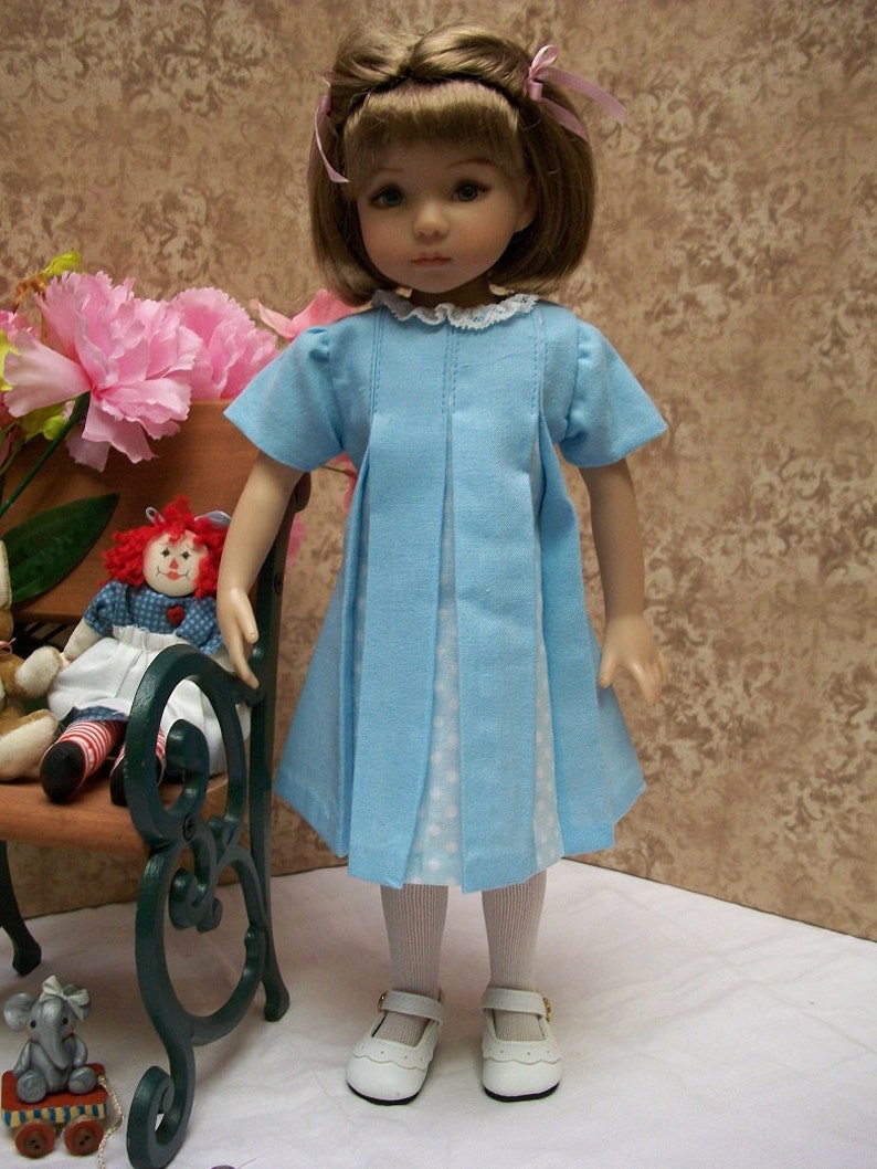 May include: A blue and white polka dot dress with a white lace collar and a pink bow in the hair. The doll is wearing white tights and white shoes.