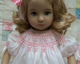 May include: A porcelain doll with blonde, curly hair and blue eyes. The doll wears a white dress with pink smocking around the neckline and pink ribbons on the sleeves. A pink bow adorns the doll's hair. The doll is a collectible item.