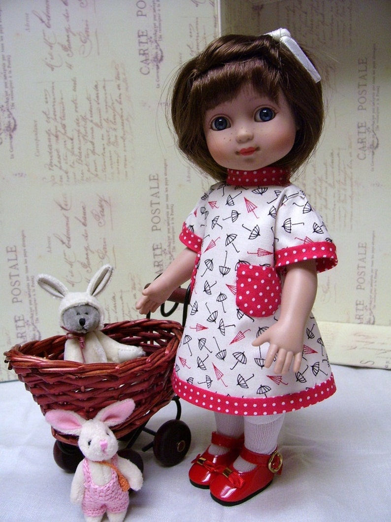 May include: A porcelain doll with brown hair wearing a white dress with red polka dots and black umbrellas printed on it. The doll is holding a wicker basket with a white bunny inside. The doll is wearing red shoes with a gold buckle.