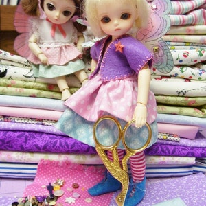 May include: Two dolls with wings, one wearing a pink dress and the other a purple jacket and pink dress, are sitting on a stack of colorful fabric. A pair of gold scissors is in front of the dolls.