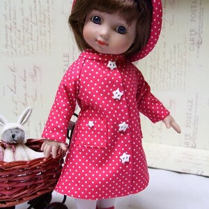 May include: A red and white polka dot raincoat with a hood, worn by a doll with brown hair and blue eyes. The doll is wearing red shoes and white tights. The doll is holding a wicker basket with a white bunny inside.