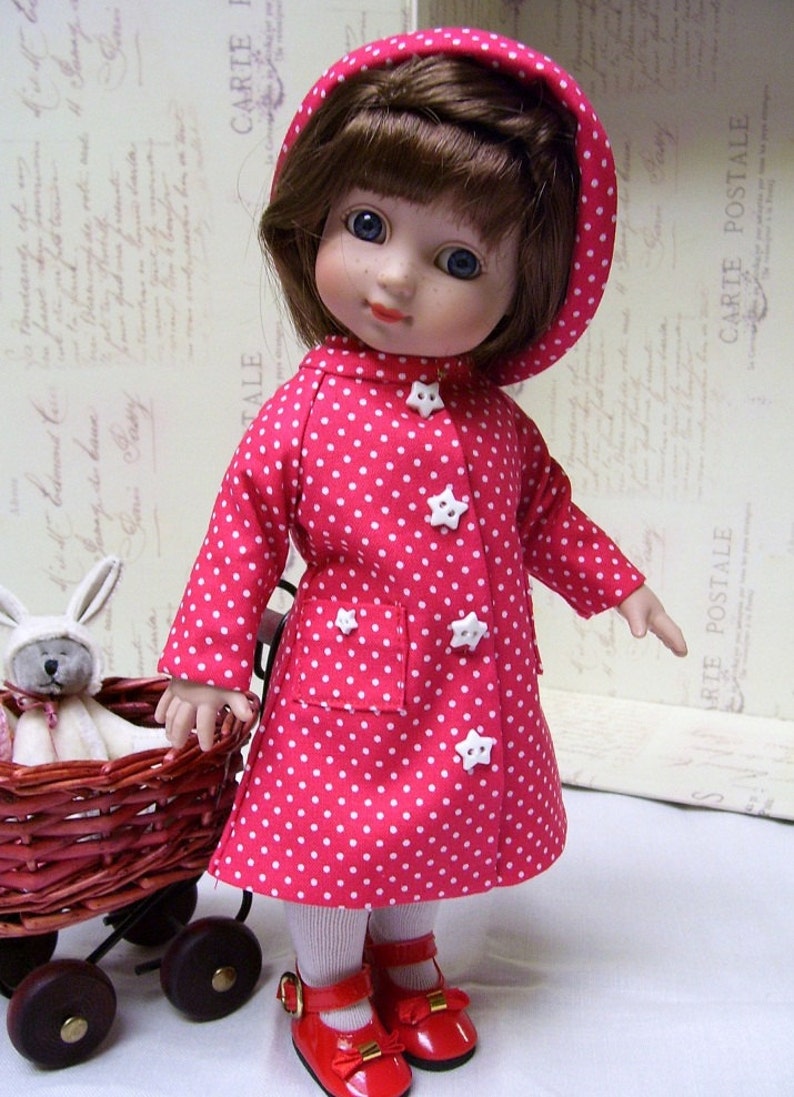 May include: A red and white polka dot raincoat with a hood, worn by a doll with brown hair and blue eyes. The doll is wearing red shoes and white tights. The doll is holding a wicker basket with a white bunny inside.
