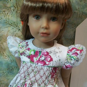 May include: A doll wearing a white smocked dress with a floral print. The dress has a gathered neckline and short sleeves with lace trim. The doll has brown hair and blue eyes.