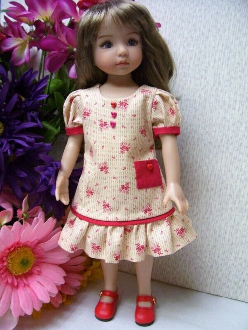 May include: A doll wearing a cream-colored dress with a red floral pattern, red trim, and a red pocket. The dress has short, puffed sleeves and a ruffled hem. The doll is wearing red shoes.