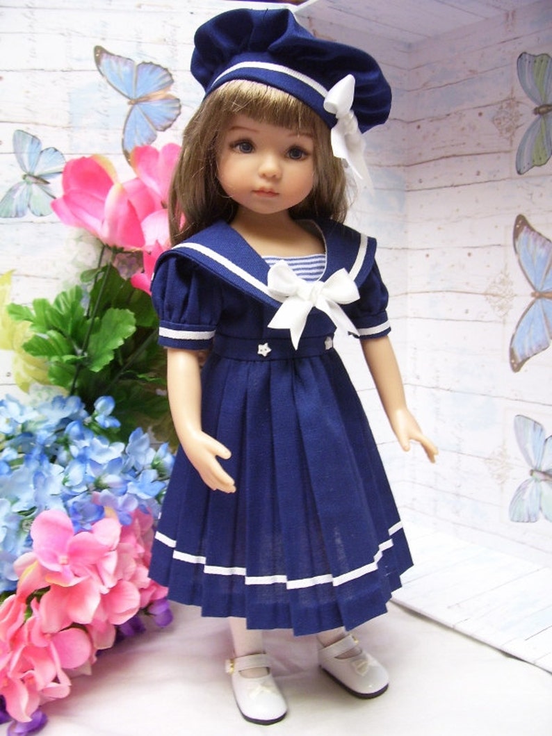 May include: A blue and white sailor dress with a white collar and a white bow. The dress has a pleated skirt and white trim. The doll is wearing a blue and white sailor hat.