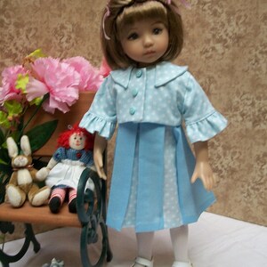May include: A doll wearing a blue and white polka dot dress with a matching jacket. The doll has brown hair and blue eyes. The doll is sitting on a bench with a red and white doll.