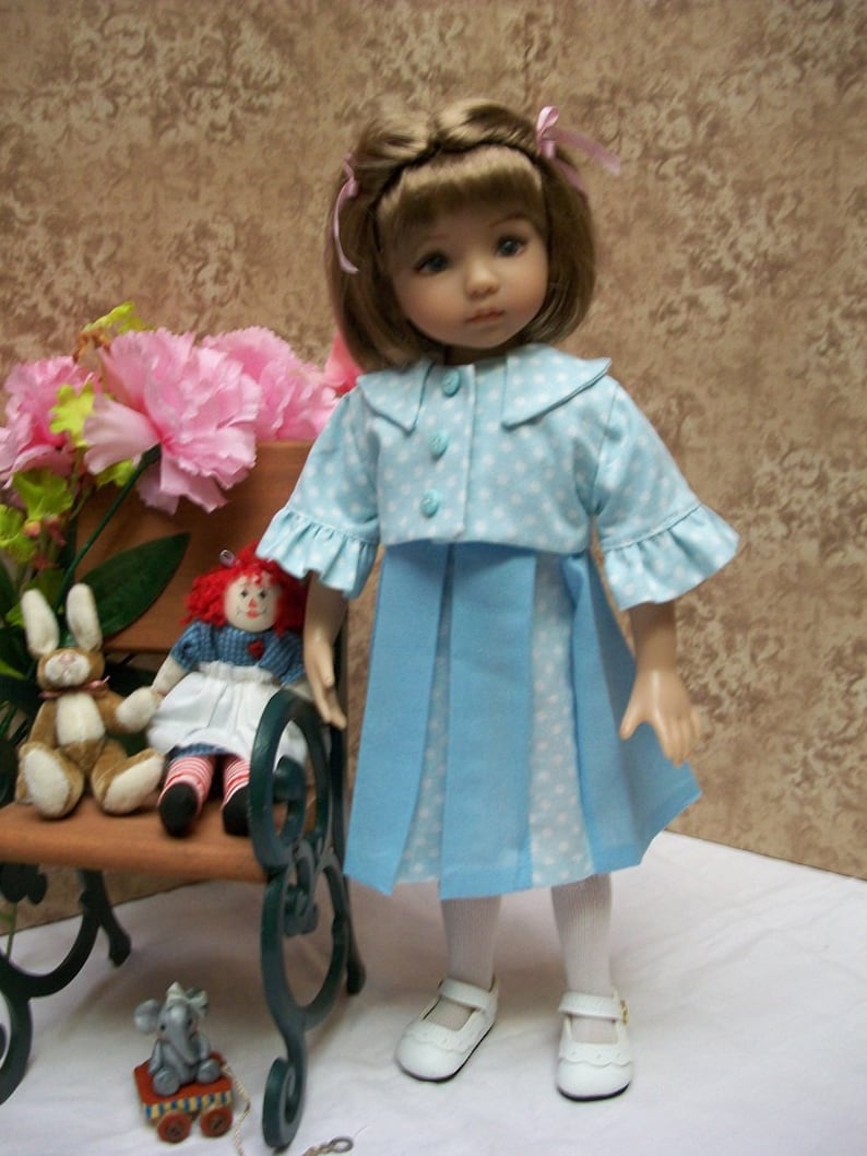 May include: A doll wearing a blue and white polka dot dress with a matching jacket. The doll has brown hair and blue eyes. The doll is sitting on a bench with a red and white doll.
