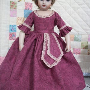 May include: A vintage porcelain doll wearing a long, magenta dress with a paisley pattern. The dress features short sleeves with ruffled cuffs and a lace trim. The doll has blonde hair and is posed in front of a patchwork quilt.