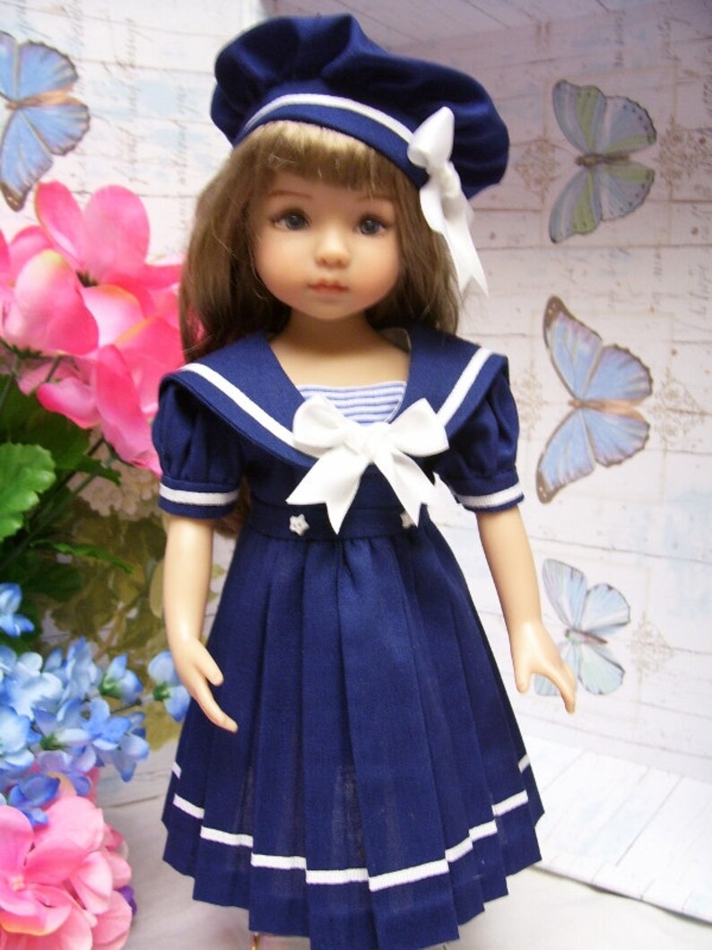 May include: A blue and white sailor dress with a white bow and a blue and white sailor hat. The dress has a pleated skirt and white trim. The doll is wearing the dress and hat.